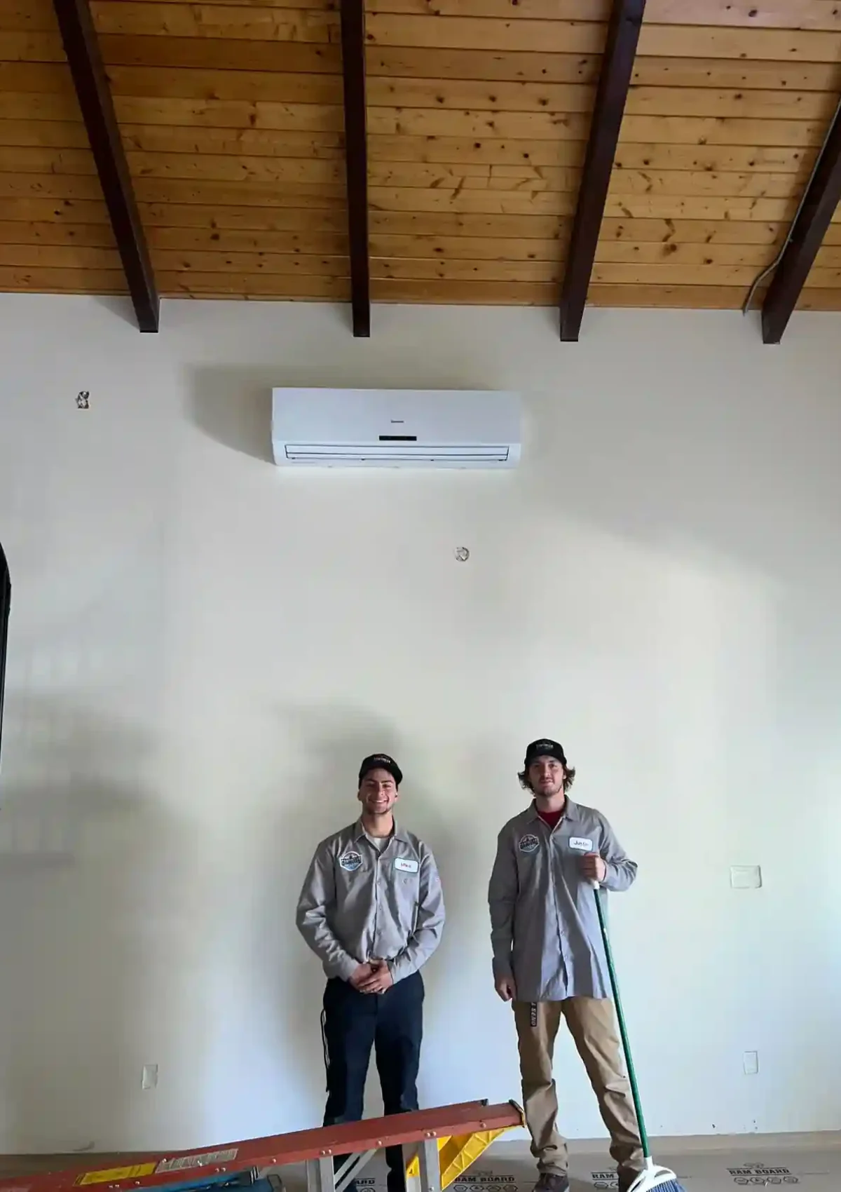 Two male HVAC contractors stand against a wall under a stand alone air conditioning unit.